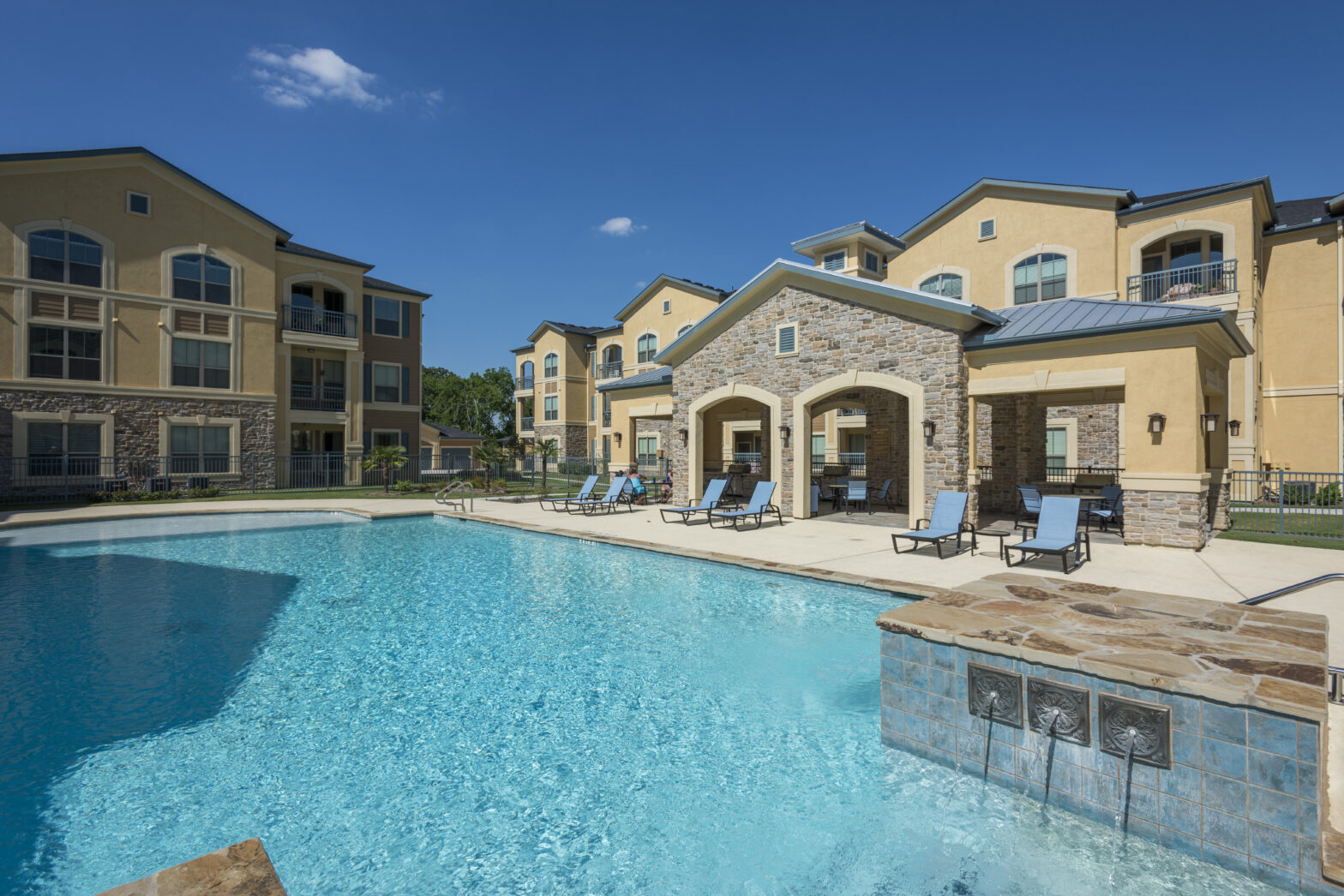 Photo of a swimming pool in front of a three-story multi-family apartment complex and clubhouse with stone facade.
